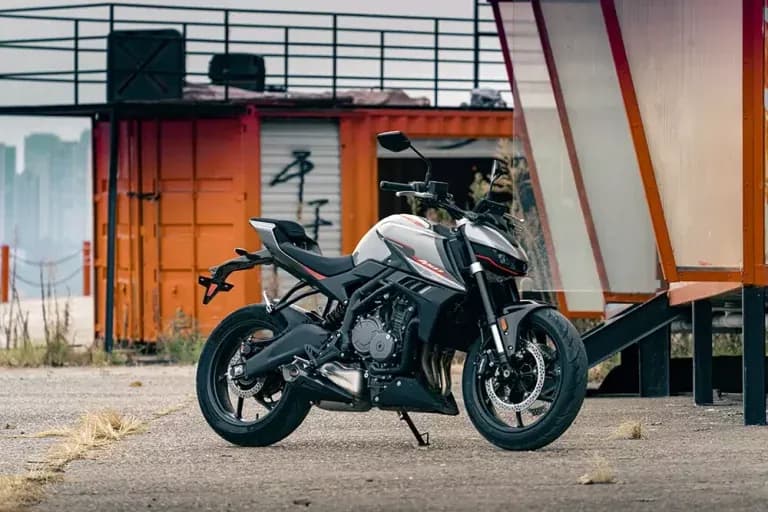 Motocicleta KOVE naked blanca y negra con detalles rojos, estacionada sobre grava frente a contenedores industriales naranjas
