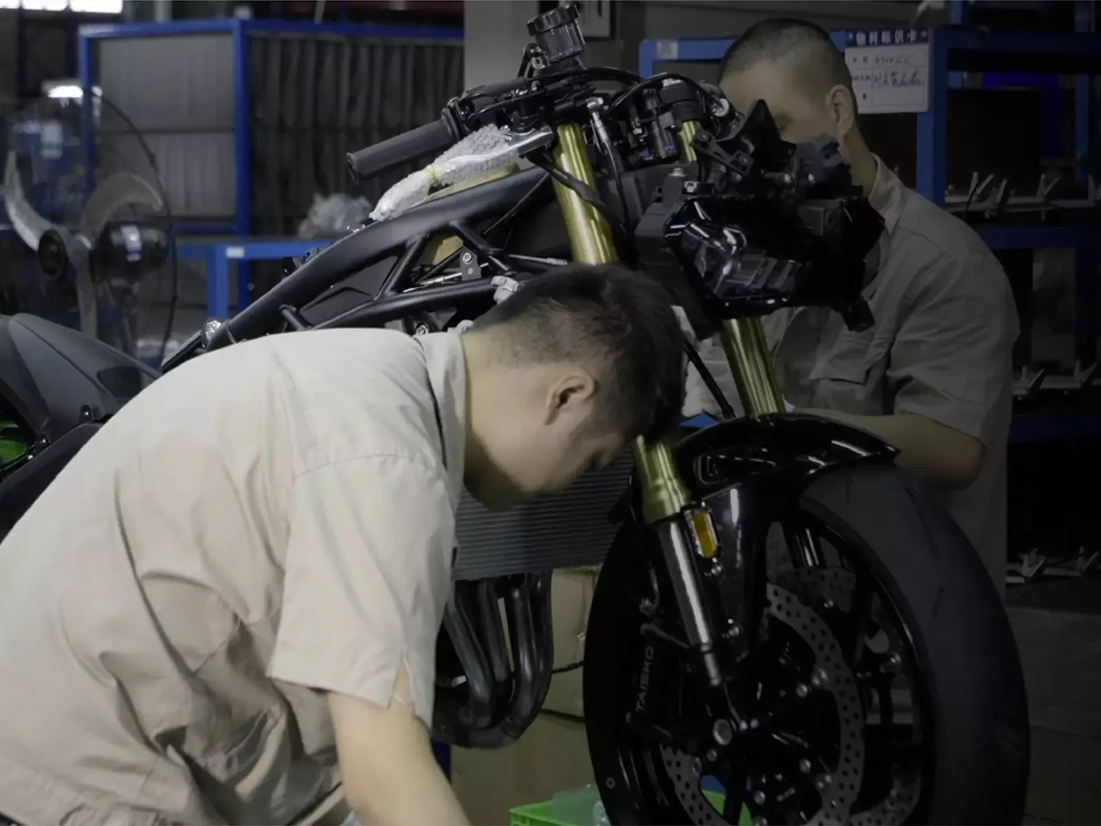 Dos operarios ensamblan una motocicleta KOVE, todavía sin carenado, en la línea de montaje de la fábrica de KOVE: se ven la horquilla dorada, el disco de freno delantero y el chasis al desnudo.